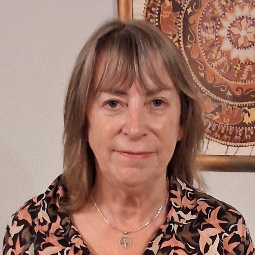 A profile photo of Diana Jackson. She's looking into the camera, with dark grey hair and a small smile. She's wearing a brown shirt with in flight birds in a rotating pattern. In the back is an Indian style tapestry.